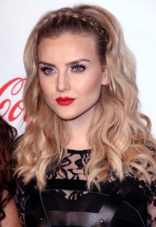 Perrie is at a red carpet event. She is not smiling in the photo. She has on big eyelashes and a red lip. Her hair is down and wavy and in a braided headband. 